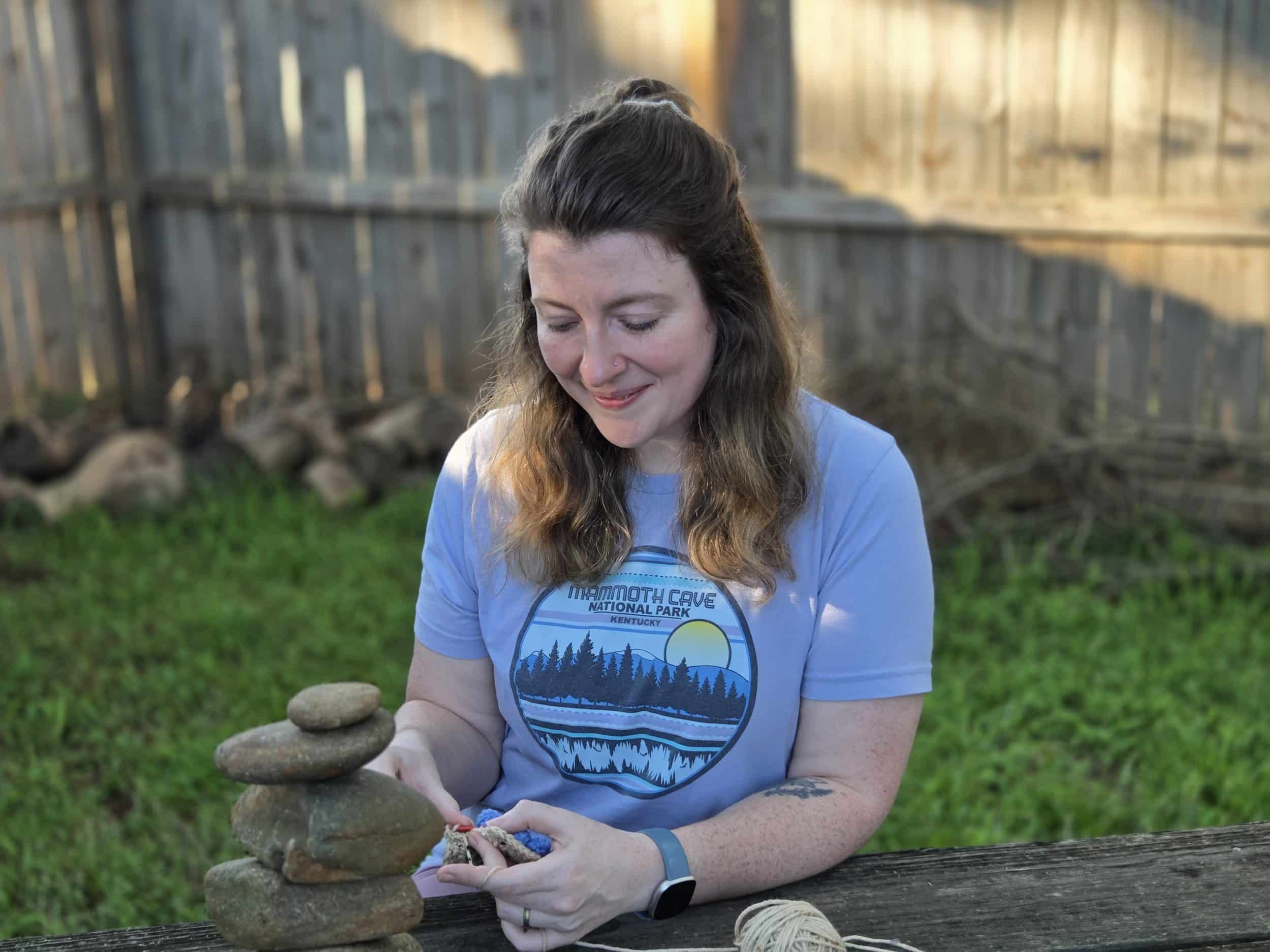 A woman crocheting outside in golden evening light, sitting at a wooden fence with a stack of balanced stones beside her and green grass in the background, representing mindful crochet and grounding in nature.