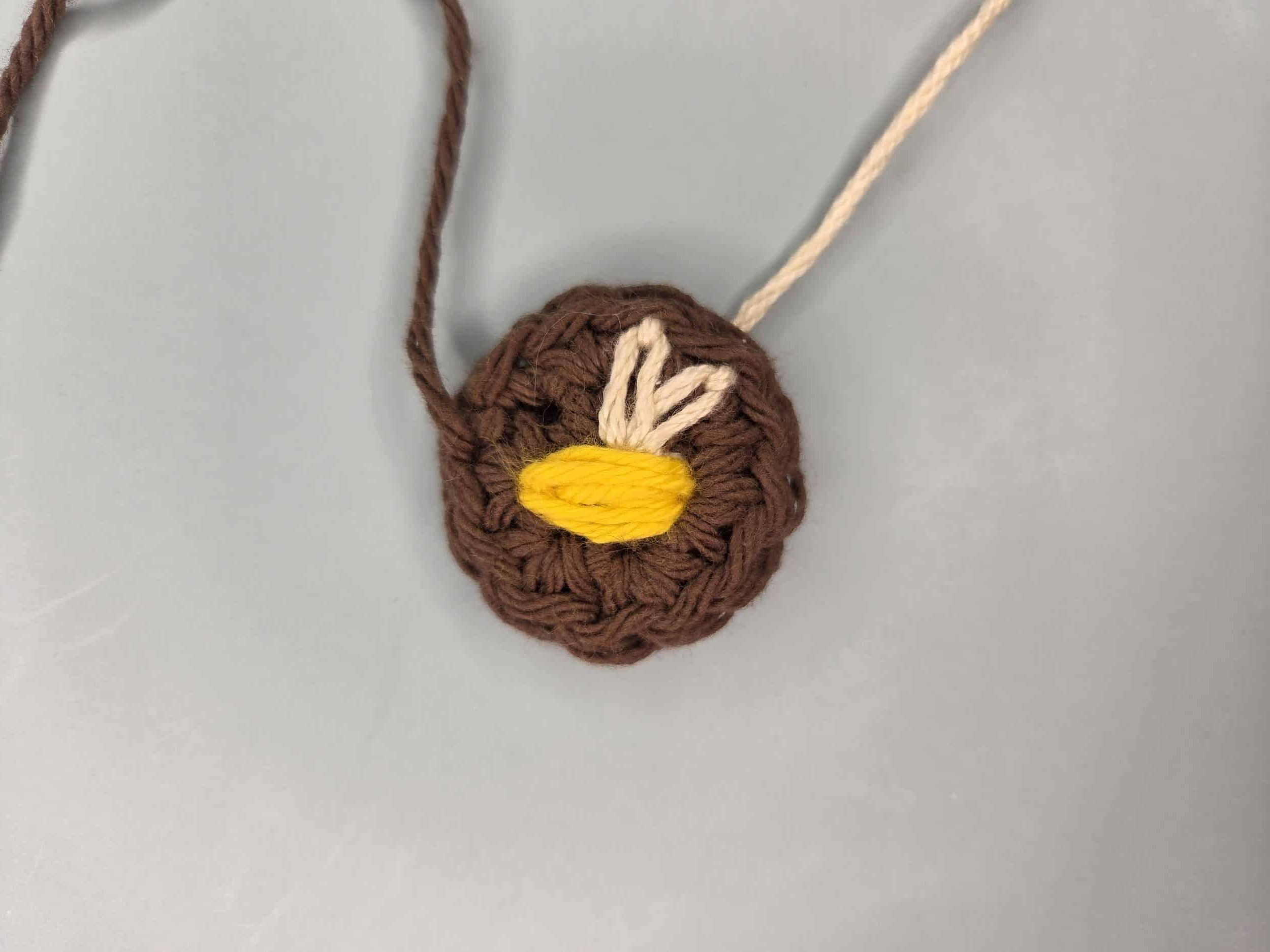 Brown crochet circle with yellow embroidered bee body and two white lazy daisy stitch wings fanning upward from the center magic ring.
