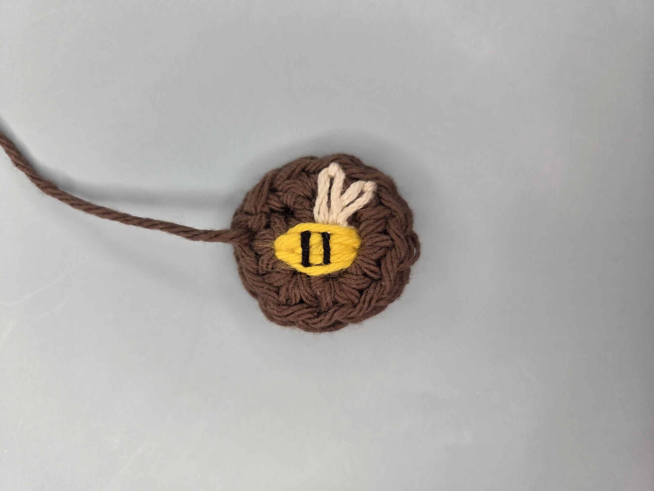 Completed embroidered bee on brown crochet circle with yellow satin stitch body, white lazy daisy wings, and two black vertical stripes across the body.