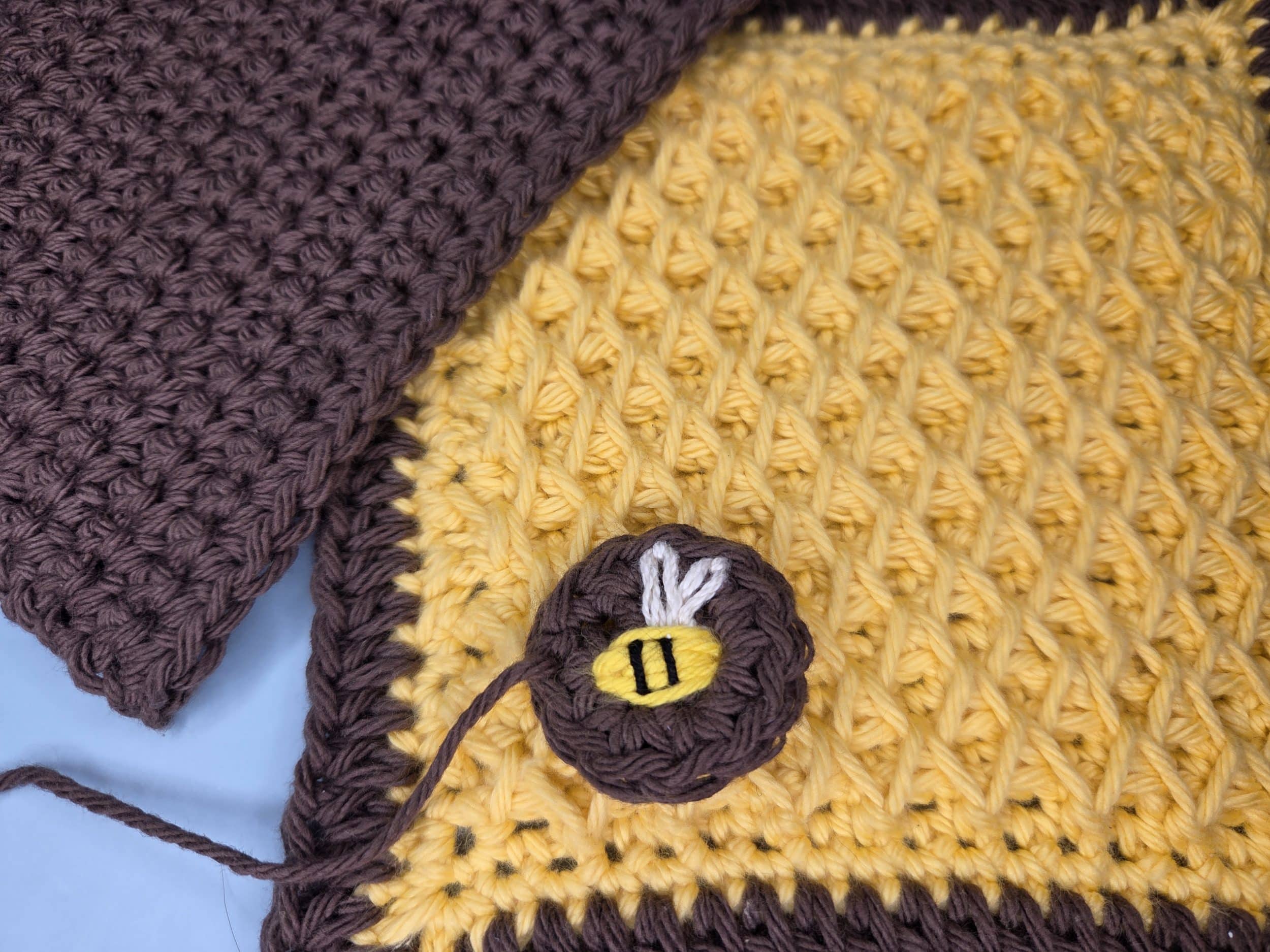 Crocheted yellow smock stitch hot pad square with brown border, brown single crochet back square, and small embroidered bee circle laid out before assembly.