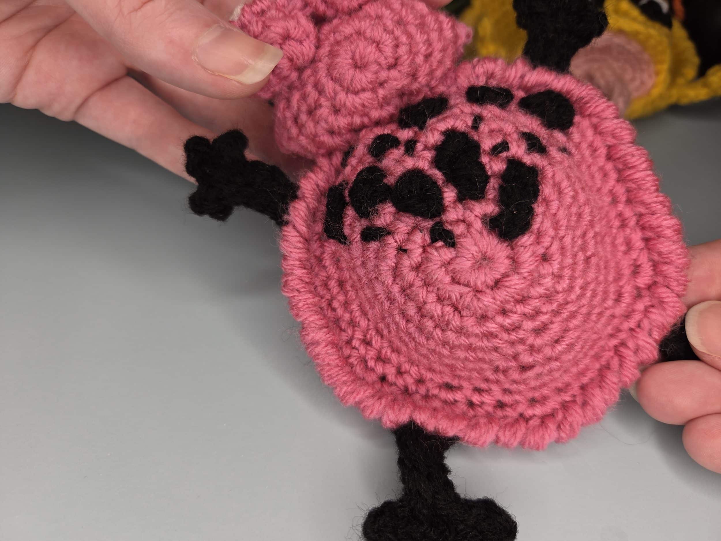 A hand holds a pink crochet circle, which forms the body of a frog, showing stitched-on black yarn spots. The body features a neat reverse single crochet, or crab stitch, border. The black yarn legs and another out-of-focus crochet item are in the background.