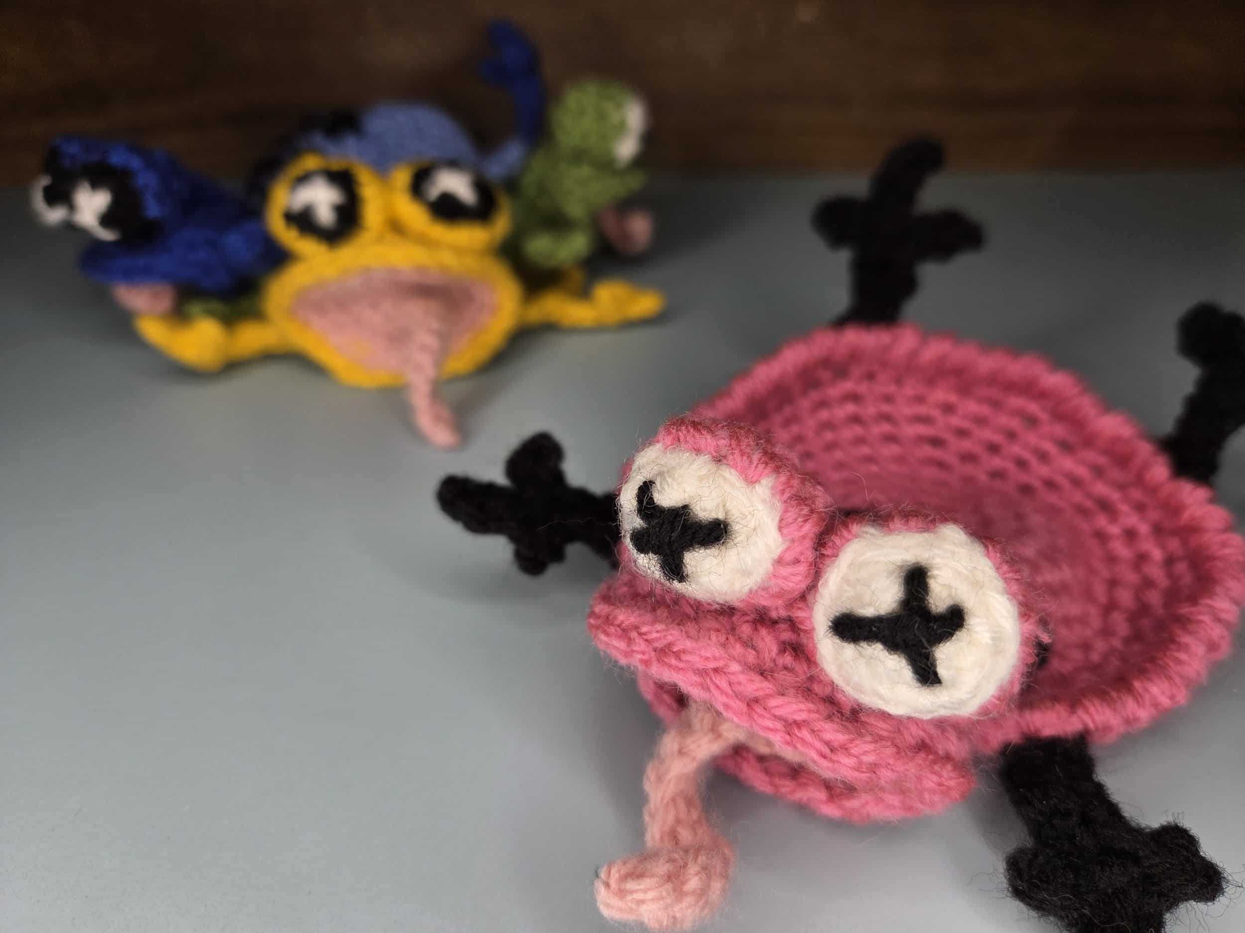 A close-up, foreground view of a finished pink crochet frog coaster with two prominent white and black "X" pupils, a pink tongue-like piece sticking out from its mouth, and black legs. In the softly blurred background, other completed multi-colored frog coasters, including a yellow one and a blue one, are visible.
