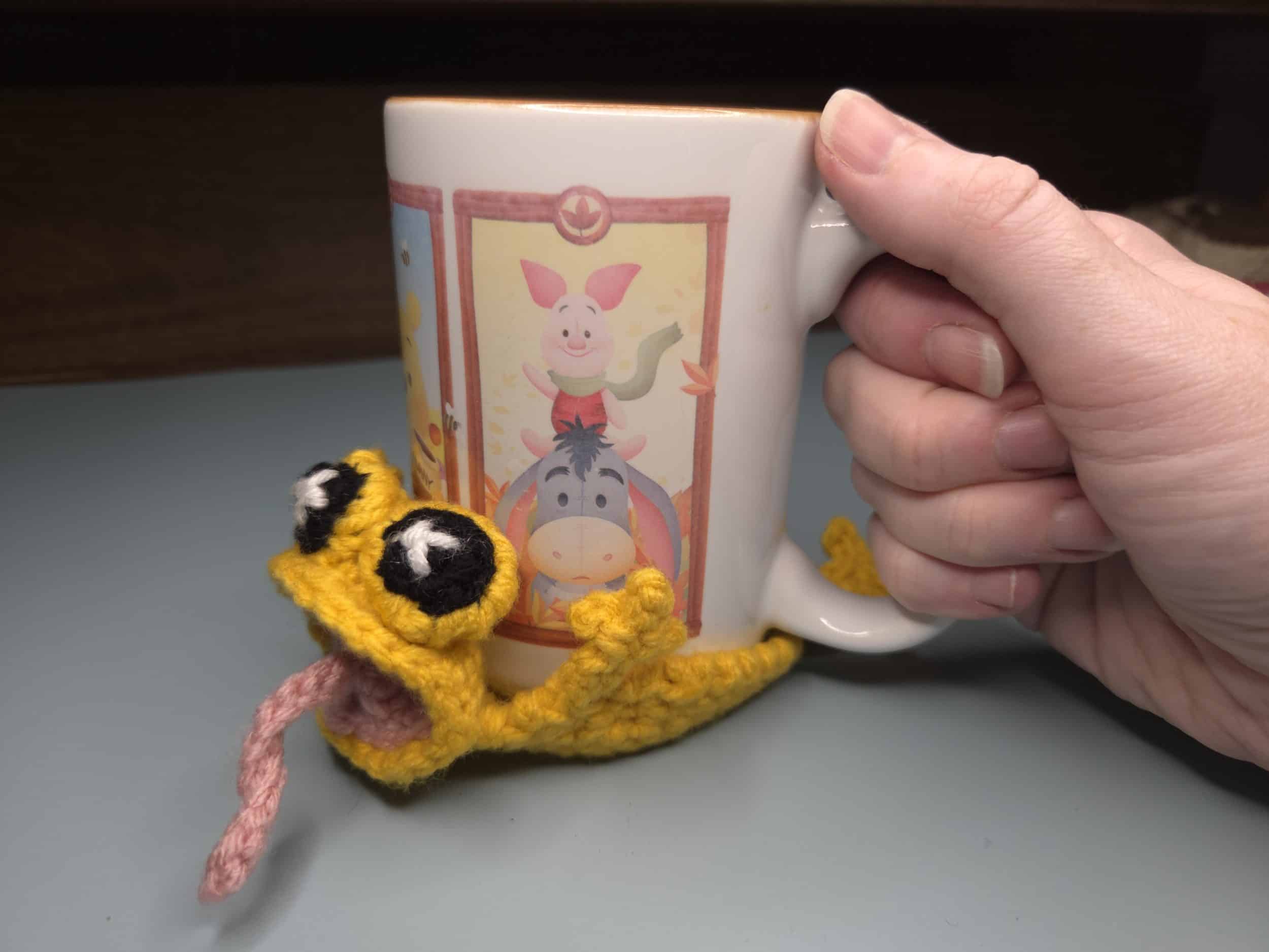 Yellow crochet frog pop coaster holding a Winnie the Pooh coffee mug, showing the concave body shape in use with legs and pink tongue visible.