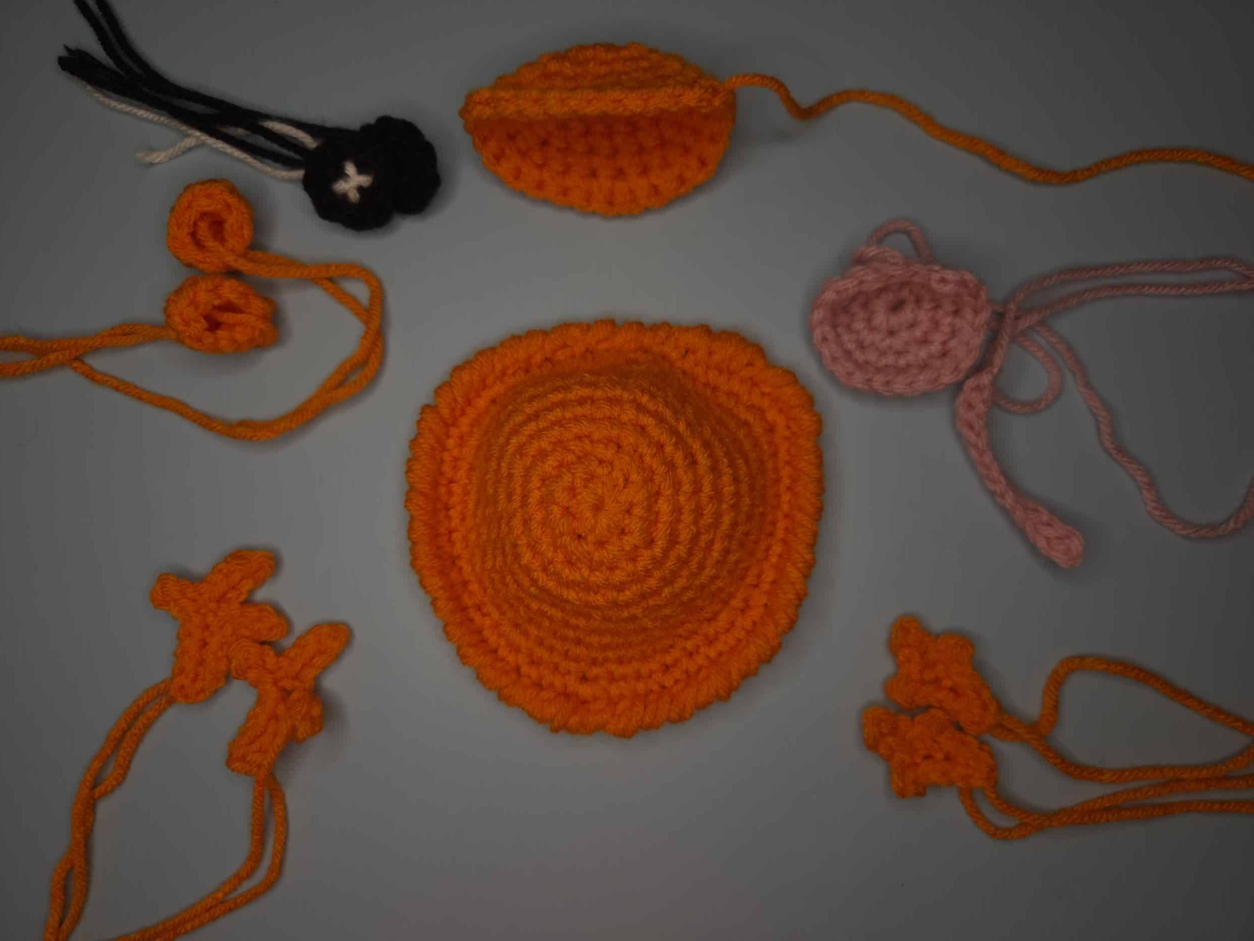 A flat-lay photograph on a neutral grey background showing all the disassembled parts of an orange crochet frog coaster. The components include the large orange coaster body, a separate orange head piece, two orange back legs, two orange front legs, two pinkish-orange smaller pieces (eyelids), and a black yarn circle embroidered X-pupil, all with long yarn tails attached, ready to be sewn together.