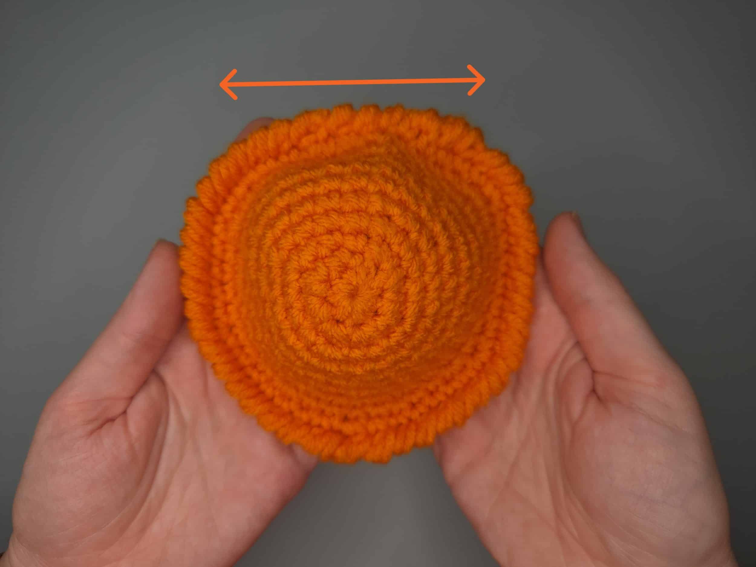Two hands holding a concave orange crochet coaster base showing the bowl shape and crab stitch edge that creates the pop coaster effect. An arrow showing the straight side.