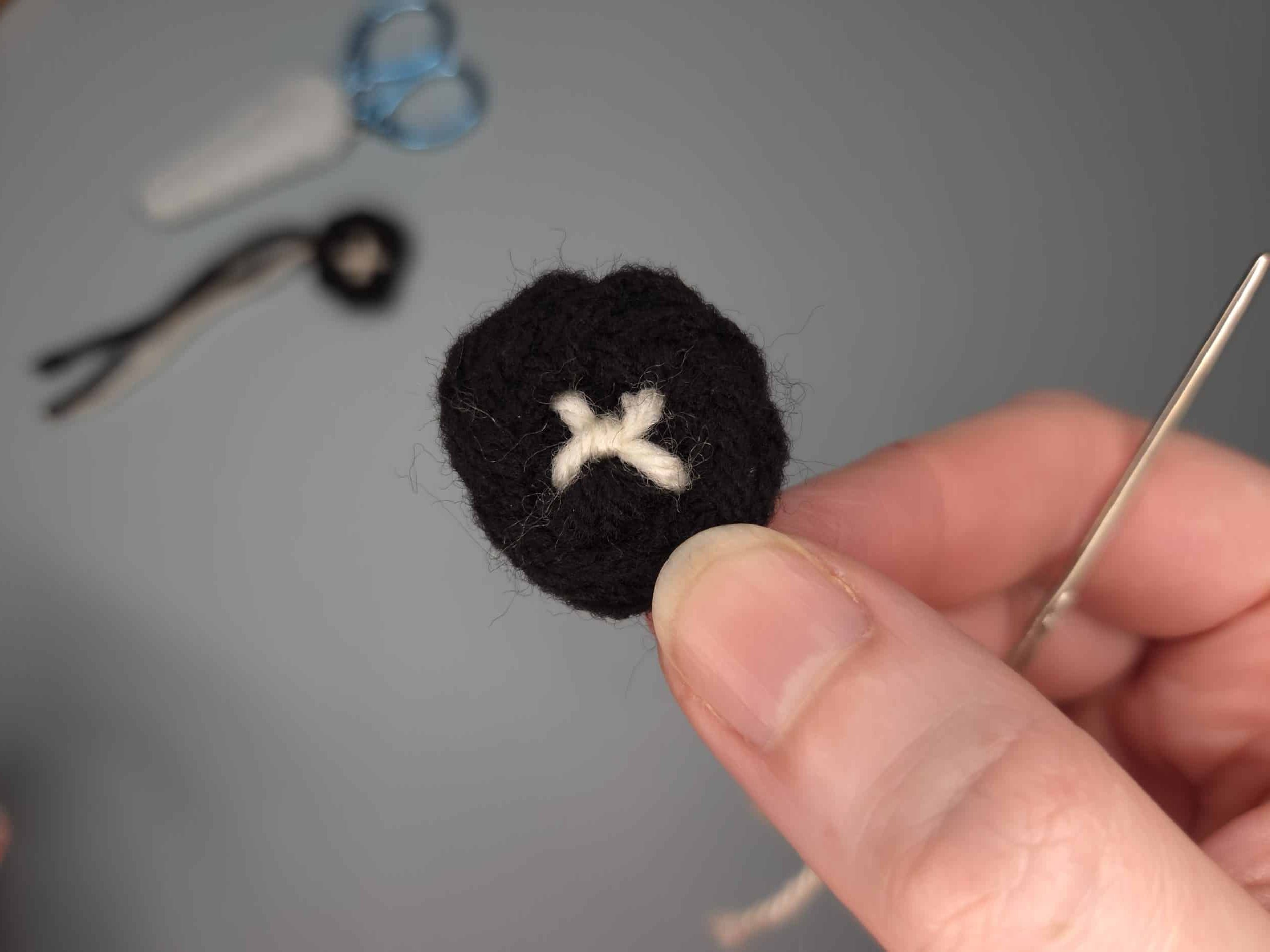 Hand holding a small black crochet circle with a white X embroidered in the center as the pupil, with scissors and a tapestry needle visible in the background.