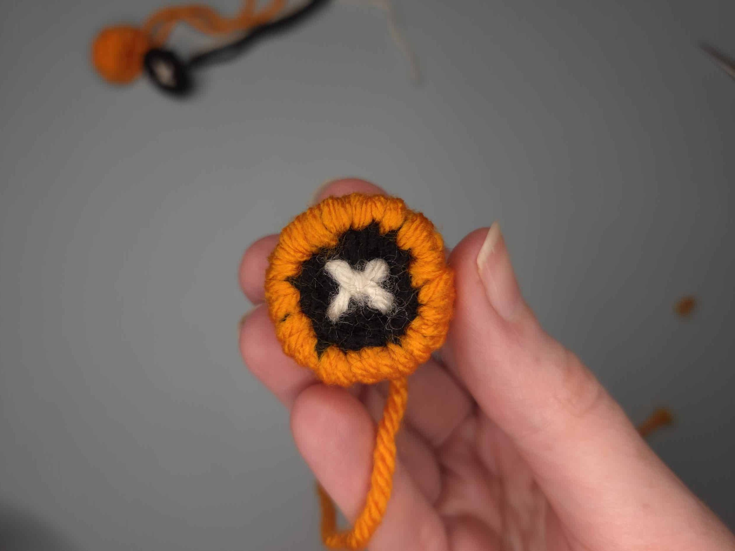 Hand holding a completed crochet frog eye showing orange outer ring, black center, and white X embroidered pupil, with sewing tail attached.